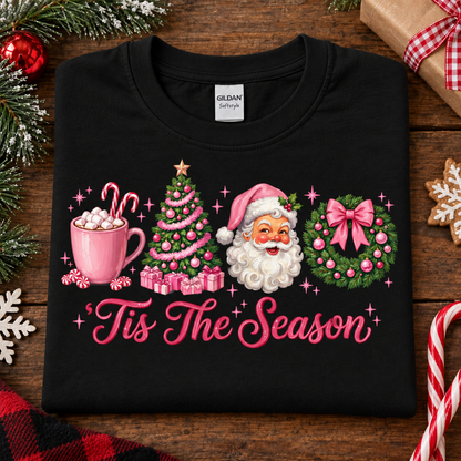 A black Gildan Comfortwear t-shirt with a colorful Christmas graphic reading 'Tis The Season'. The graphic features a pink mug with hot chocolate and marshmallows, a pink Christmas tree with presents, Santa Claus, and a pink and green wreath.