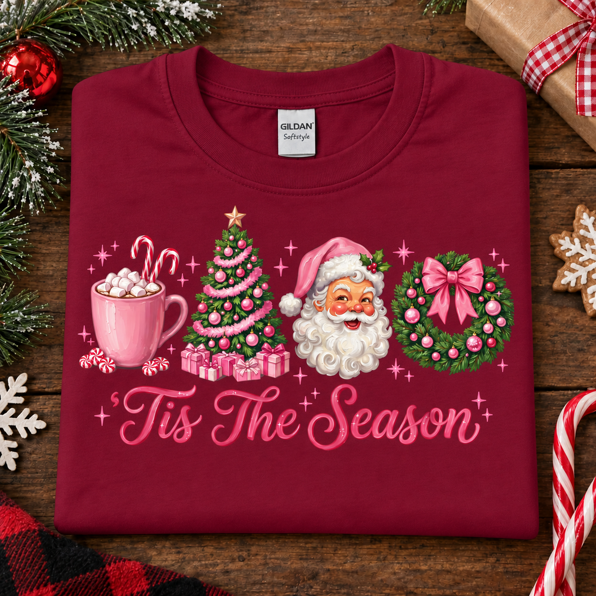 A folded cardinal red Gildan Comfortware t-shirt with a colorful Christmas graphic that reads 'Tis The Season'. The graphic features a pink mug with marshmallows, a pink Christmas tree, Santa Claus, and a pink bow-wrapped wreath.