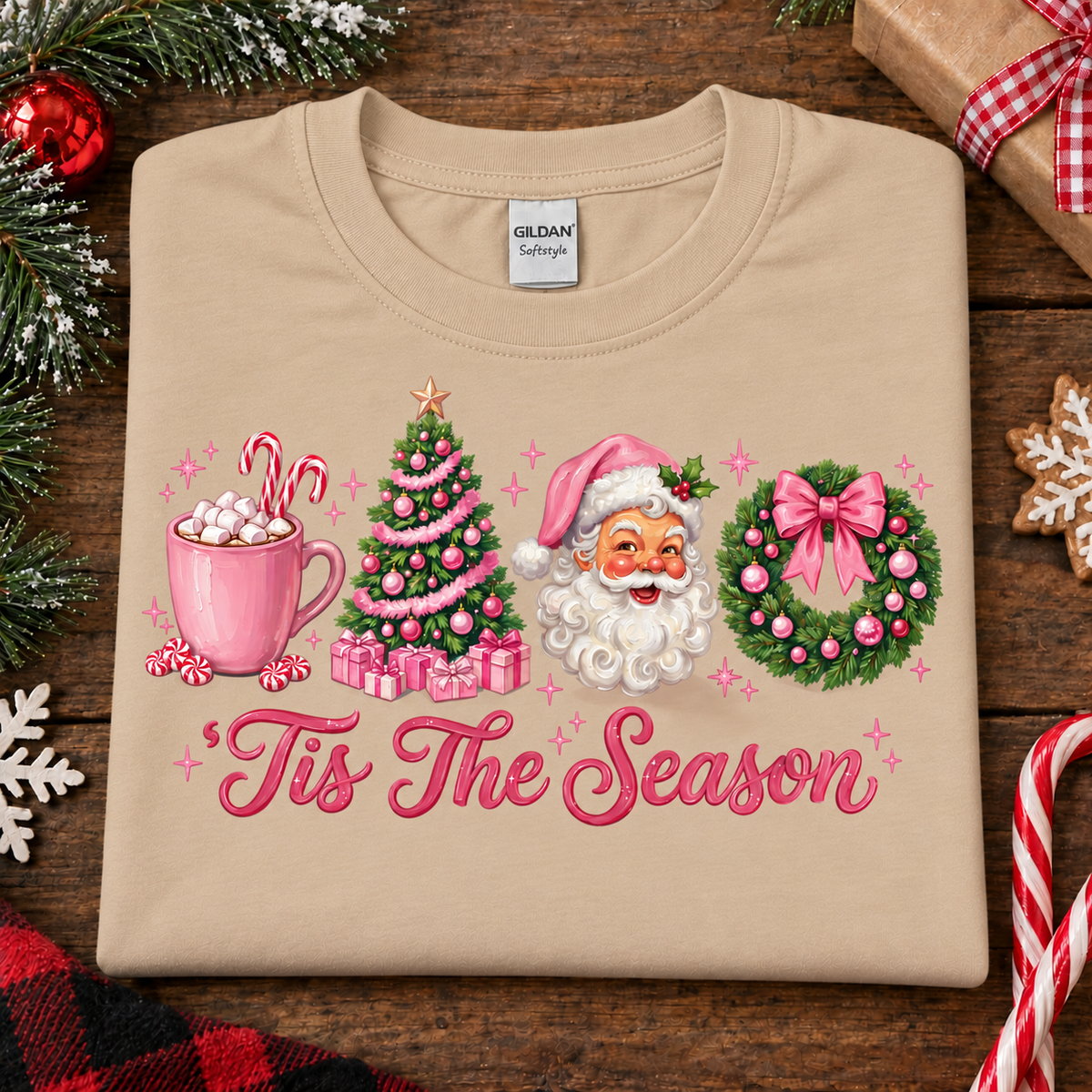 A sandcolored Gildan Softstyle t-shirt with a festive Christmas graphic, laid flat on a rustic wooden background with holiday decorations. The graphic features a pink mug with hot cocoa and marshmallows, a decorated Christmas tree, Santa Claus, a pink bow on a green wreath, and the text 'Tis The Season' in pink cursive.