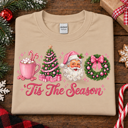 A sandcolored Gildan Softstyle t-shirt with a festive Christmas graphic, laid flat on a rustic wooden background with holiday decorations. The graphic features a pink mug with hot cocoa and marshmallows, a decorated Christmas tree, Santa Claus, a pink bow on a green wreath, and the text 'Tis The Season' in pink cursive.