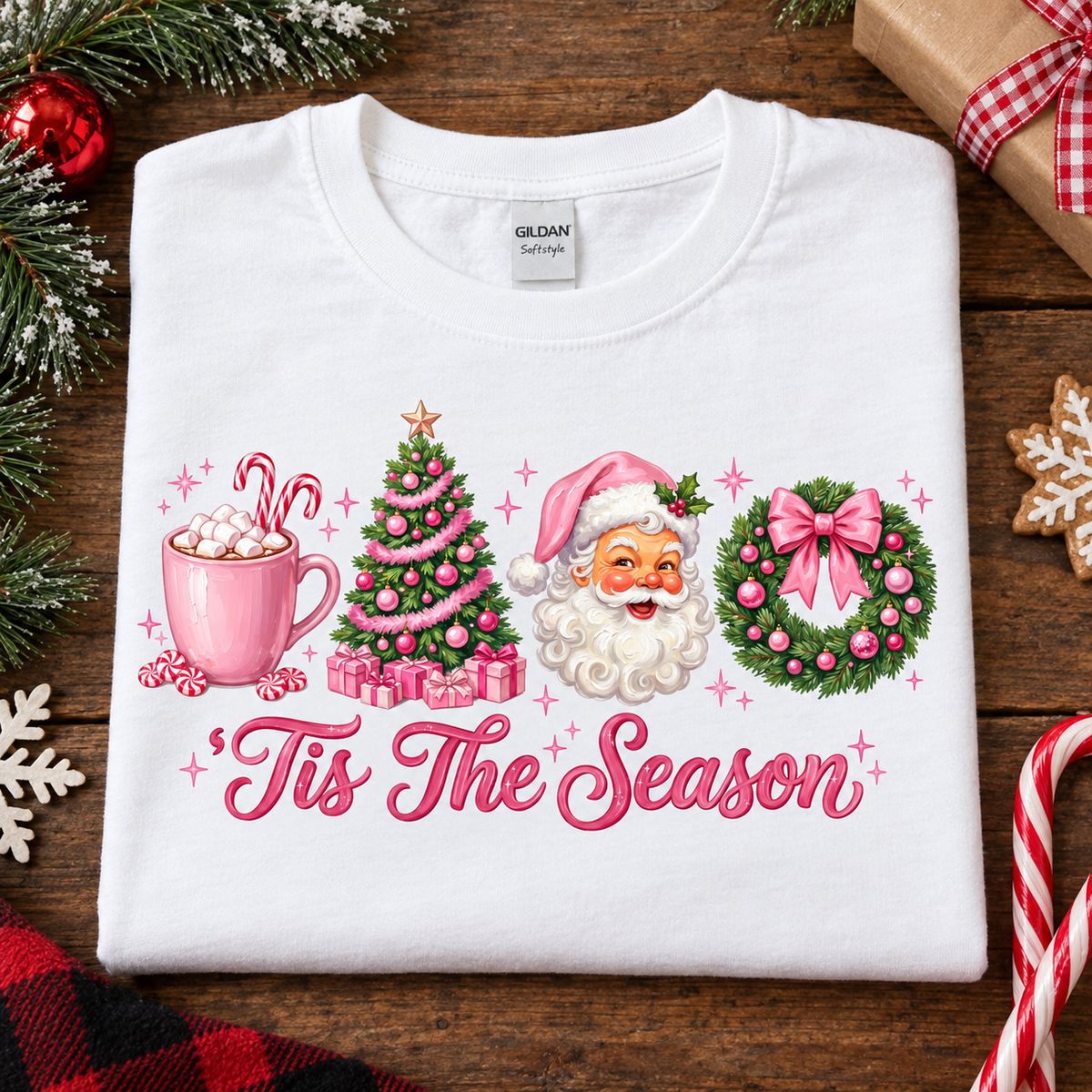 A white Gildan Softstyle crewneck t-shirt with a colorful Christmas graphic print, laid flat on a rustic wooden background decorated with holiday ornaments, pine branches, and wrapped gifts.