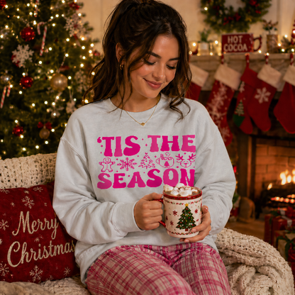 Woman wearing a 'Tis the Season ash gray sweatshirt holding a mug of hot chocolate in a festive setting with Christmas decorations.