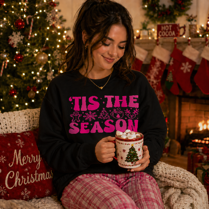 Woman in 'Tis the Season black sweatshirt holding a mug with Christmas decorations, sitting in a festive living room.