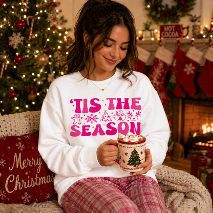 Woman wearing a 'Tis the Season sweater holding a mug in a festive setting with Christmas decorations.