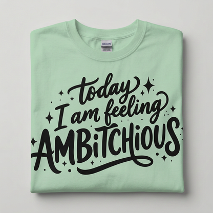 A folded mint green Gildan t-shirt with a black hand-lettered graphic that reads 'today I am feeling AMBITCHIOUS' surrounded by small star and dot accents, displayed on a neutral light gray background.