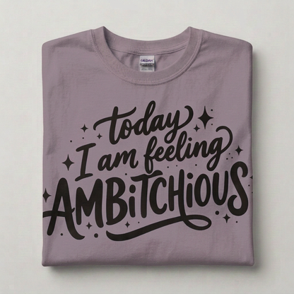 A folded paragon purple crewneck t-shirt with a black graphic text print reading 'today I am feeling AMBITIOUS' surrounded by small star doodles. The shirt is laid flat on a light gray background, showing a Gildan brand tag inside the collar.