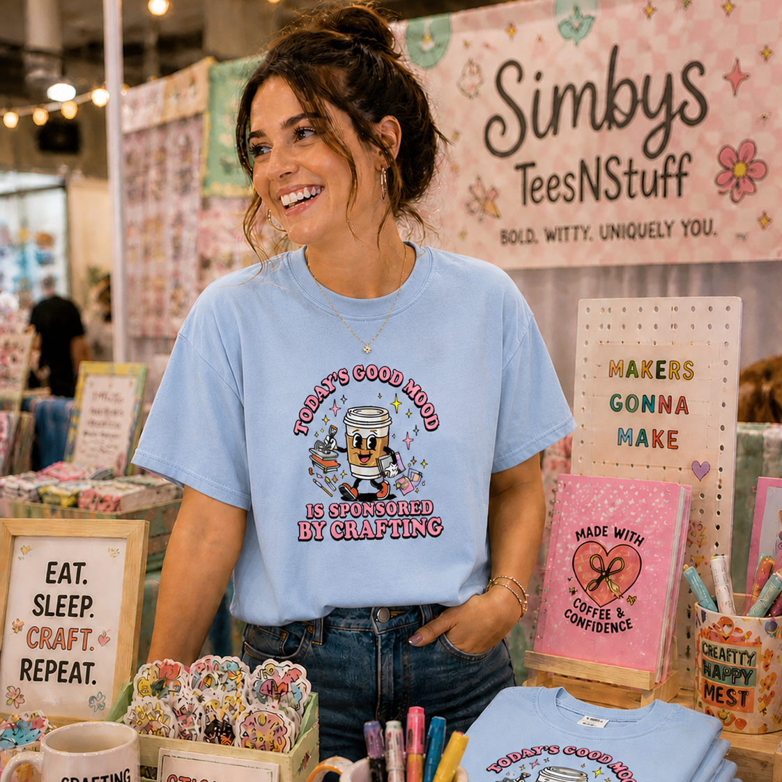 Woman wearing a blue t-shirt with a graphic design at a booth named 'Simby's Tees N Stuff'.