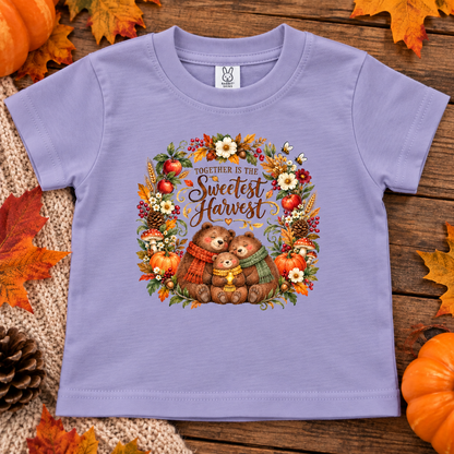 A lavender toddler t-shirt with a 'Together is the Sweetest Harvest' graphic featuring two teddy bears in autumn colors, styled on a rustic wooden background with pumpkins and fall leaves.