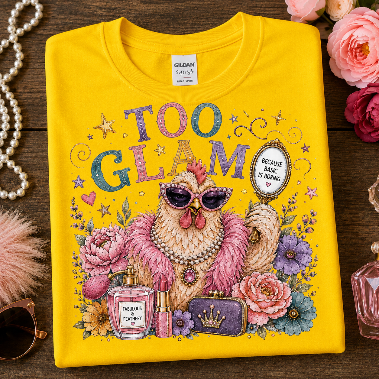 A folded daisy colored Gildan t-shirt with a colorful graphic print of a chicken in pink feathers, sunglasses, and a pearl necklace, surrounded by flowers, perfume bottles, and lipstick tubes. The shirt is styled on a dark wood background with decorative pink and red roses, a pearl strand, and a pink feather boa.