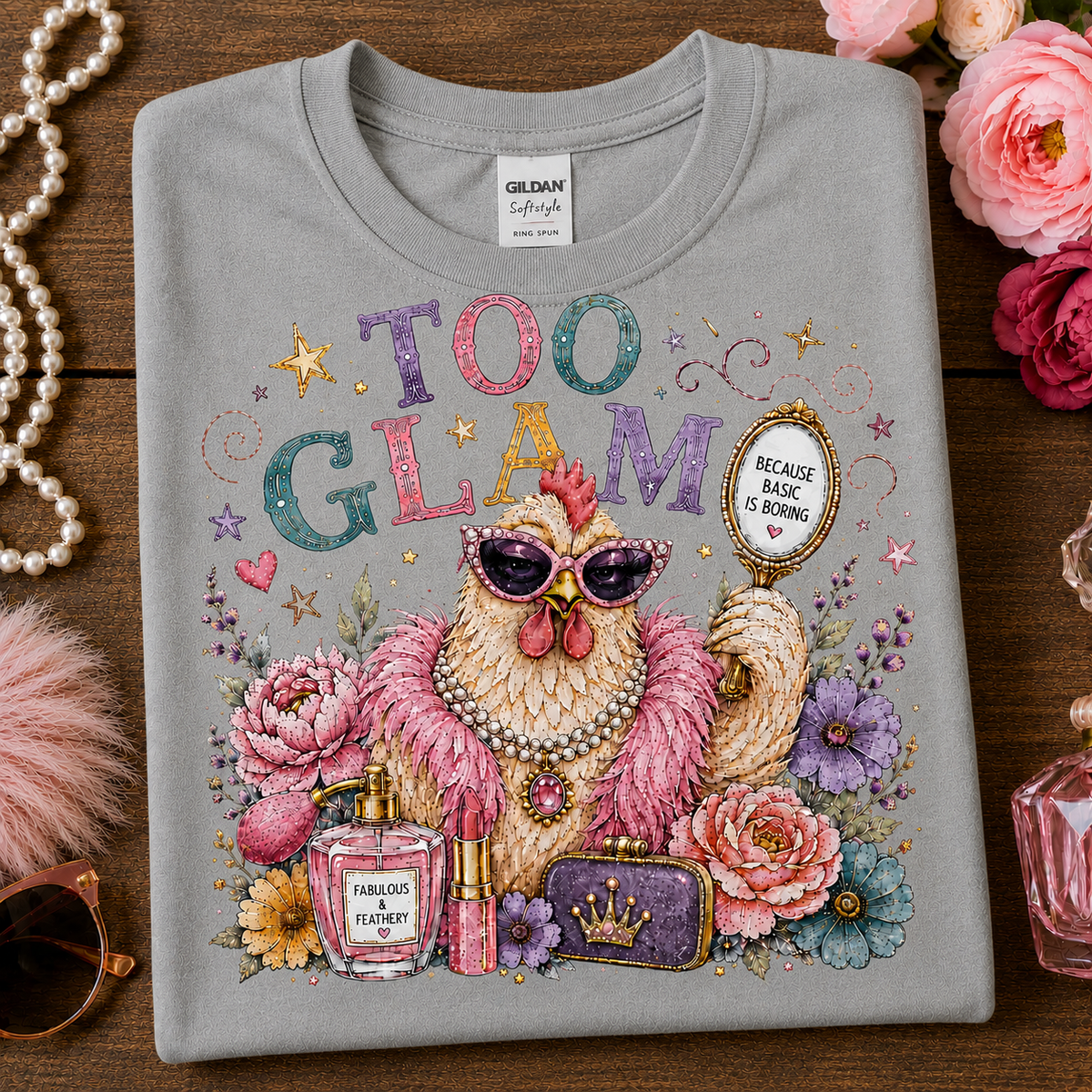 A folded sport gray t-shirt with a colorful graphic print of a fashionable chicken wearing sunglasses and a pearl necklace, surrounded by flowers, perfume bottles, and a mirror that reads 'BEIGE BLACK OR BURNING'. The shirt is displayed on a wooden surface with decorative props including pink roses, a pearl necklace, and sunglasses.
