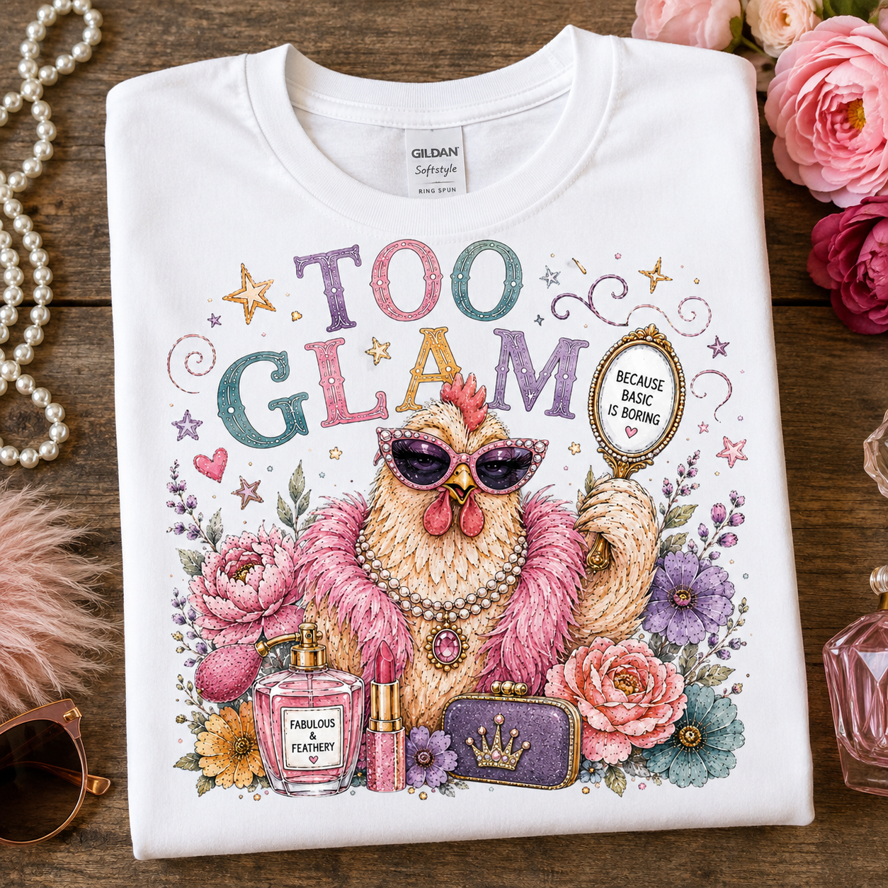 A folded white Gildan Softee T-shirt is displayed on a rustic wooden surface surrounded by pink roses, a pearl necklace, a pink feather boa, and perfume bottles. The shirt features a colorful graphic of a chicken wearing sunglasses and a pink feather boa, surrounded by flowers and cosmetics, with the text 'TOO GLAM' and a badge reading 'BECAUSE BRASH IS BORN'.