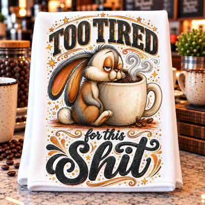 A white kitchen towel with a humorous graphic print featuring a tired brown and white rabbit sitting in a coffee mug, surrounded by the text 'TOO TIRED for this Shit' in stylized lettering. 