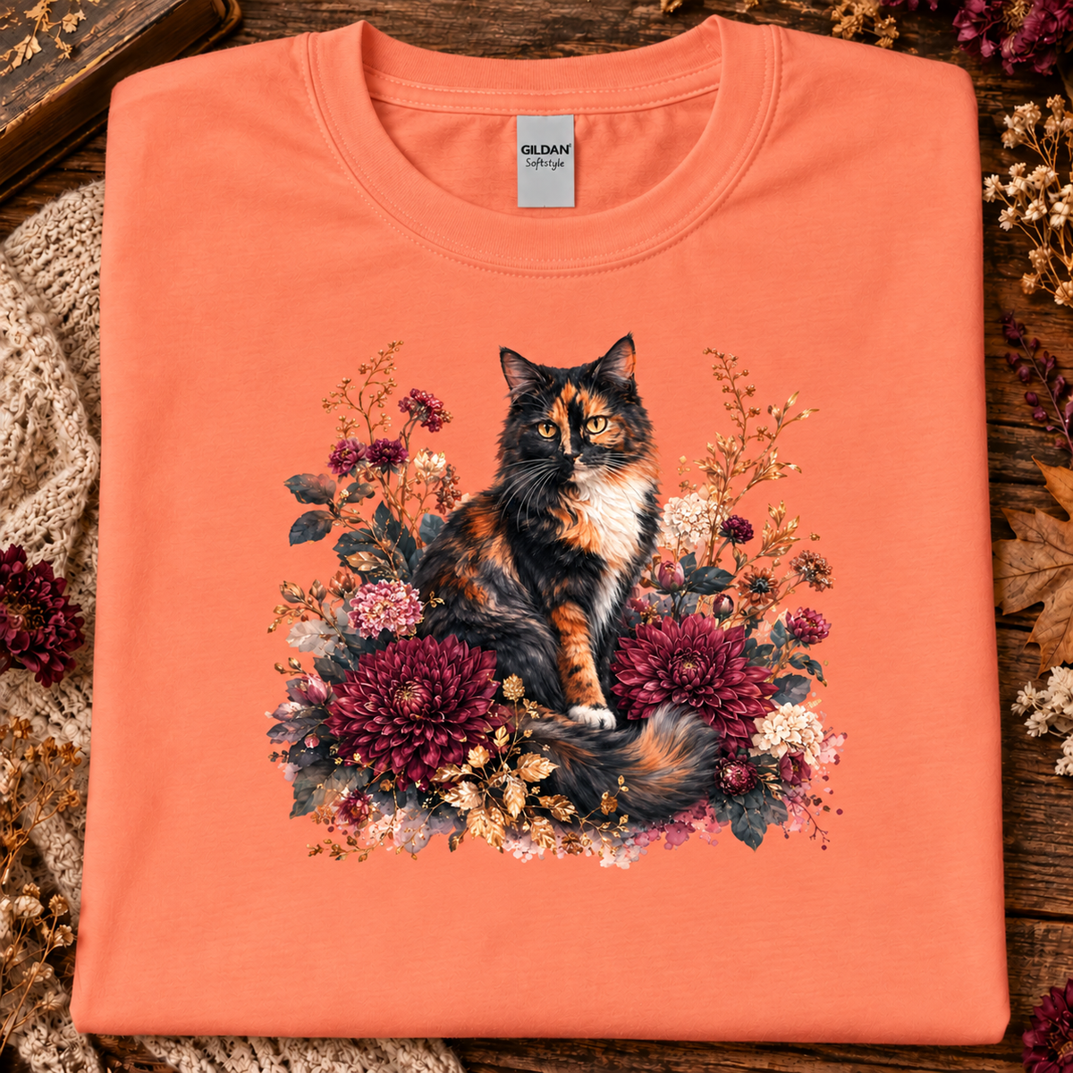 A fan texas orange colored t-shirt with a stylized graphic of a fluffy calico cat surrounded by large burgundy chrysanthemums, cream flowers, and muted foliage on a white background
