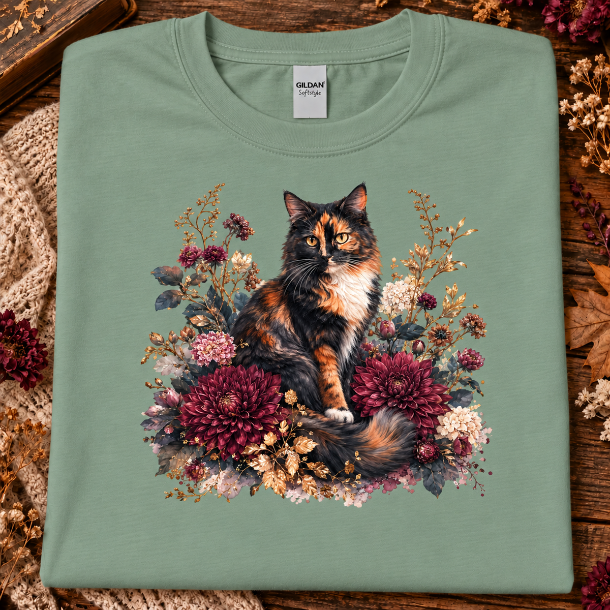 A sage colored t-shirt with a stylized graphic of a fluffy calico cat surrounded by large burgundy chrysanthemums, cream flowers, and muted foliage on a white background