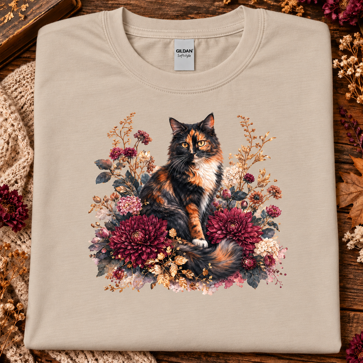 A sand colored t-shirt with a stylized graphic of a fluffy calico cat surrounded by large burgundy chrysanthemums, cream flowers, and muted foliage on a white background