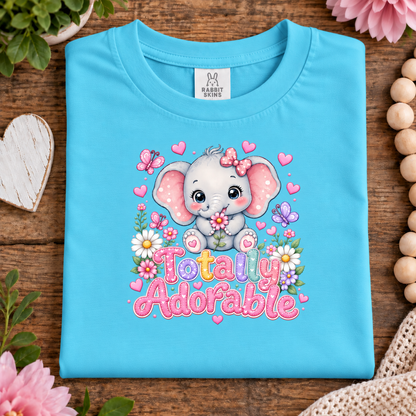 A folded aqua blue t-shirt with a cute cartoon baby elephant graphic and the text 'Totally Adorable' in pink letters, styled on a wooden surface with flowers, beads, and a heart-shaped wooden decor piece.