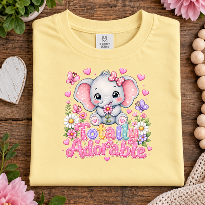 A folded butter yellow T-shirt with a colorful cartoon elephant graphic and the words 'Totally Adorable' on the front, styled on a wooden surface with flowers, beads, and a heart-shaped prop.