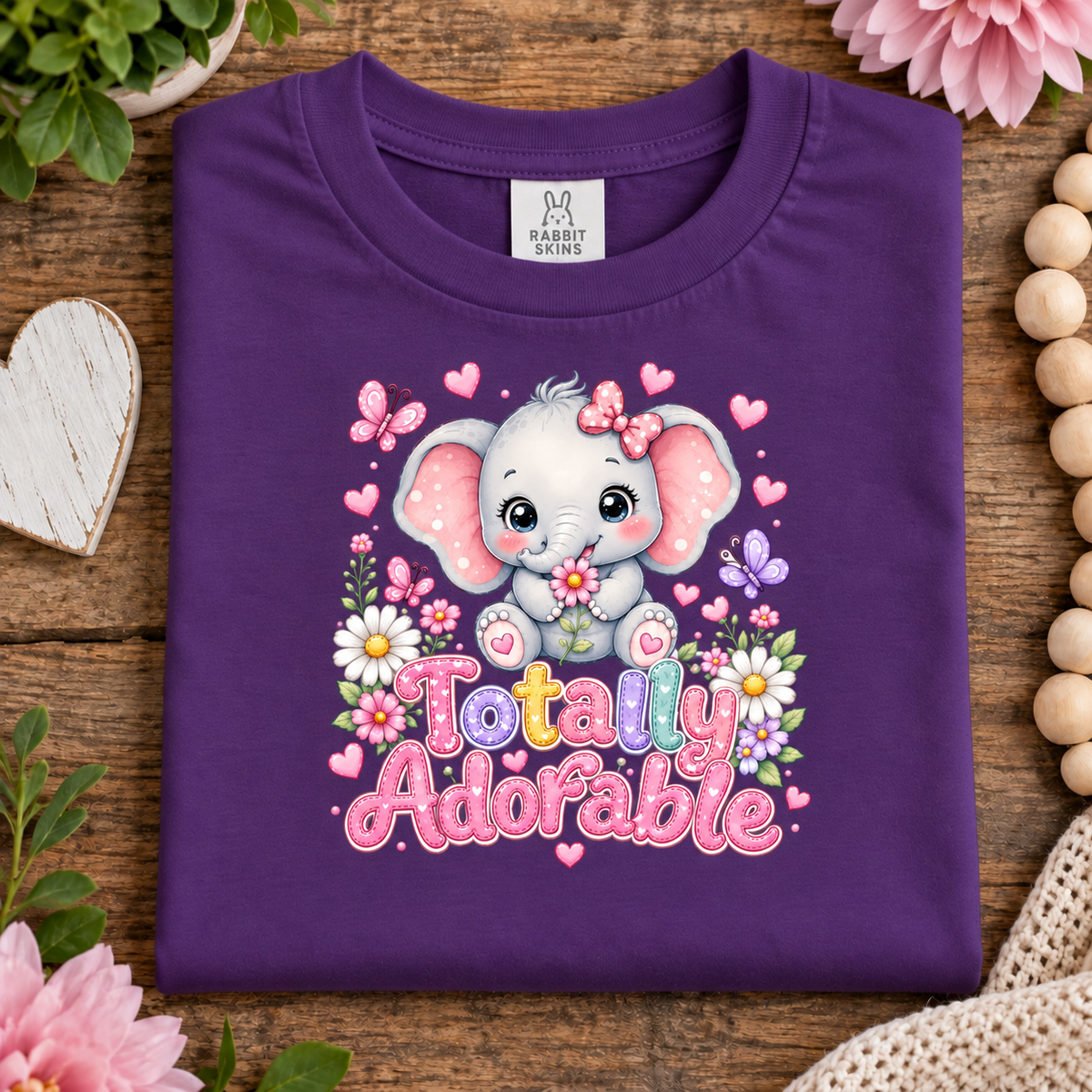 A folded purple t-shirt with a cute cartoon baby elephant graphic and the text 'Totally Adorable' in colorful letters, surrounded by pink flowers, butterflies, and hearts. The shirt is displayed on a rustic wooden background with decorative props like a heart-shaped coaster, beads, and plants.