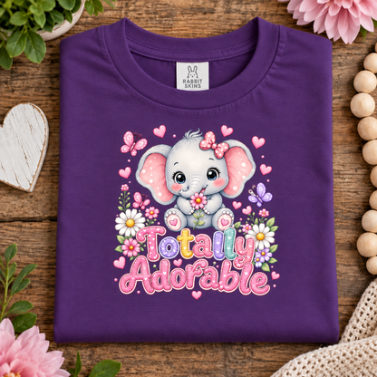A folded purple t-shirt with a cute cartoon baby elephant graphic and the text 'Totally Adorable' in colorful letters, surrounded by pink flowers, butterflies, and hearts. The shirt is displayed on a rustic wooden background with decorative props like a heart-shaped coaster, beads, and plants.
