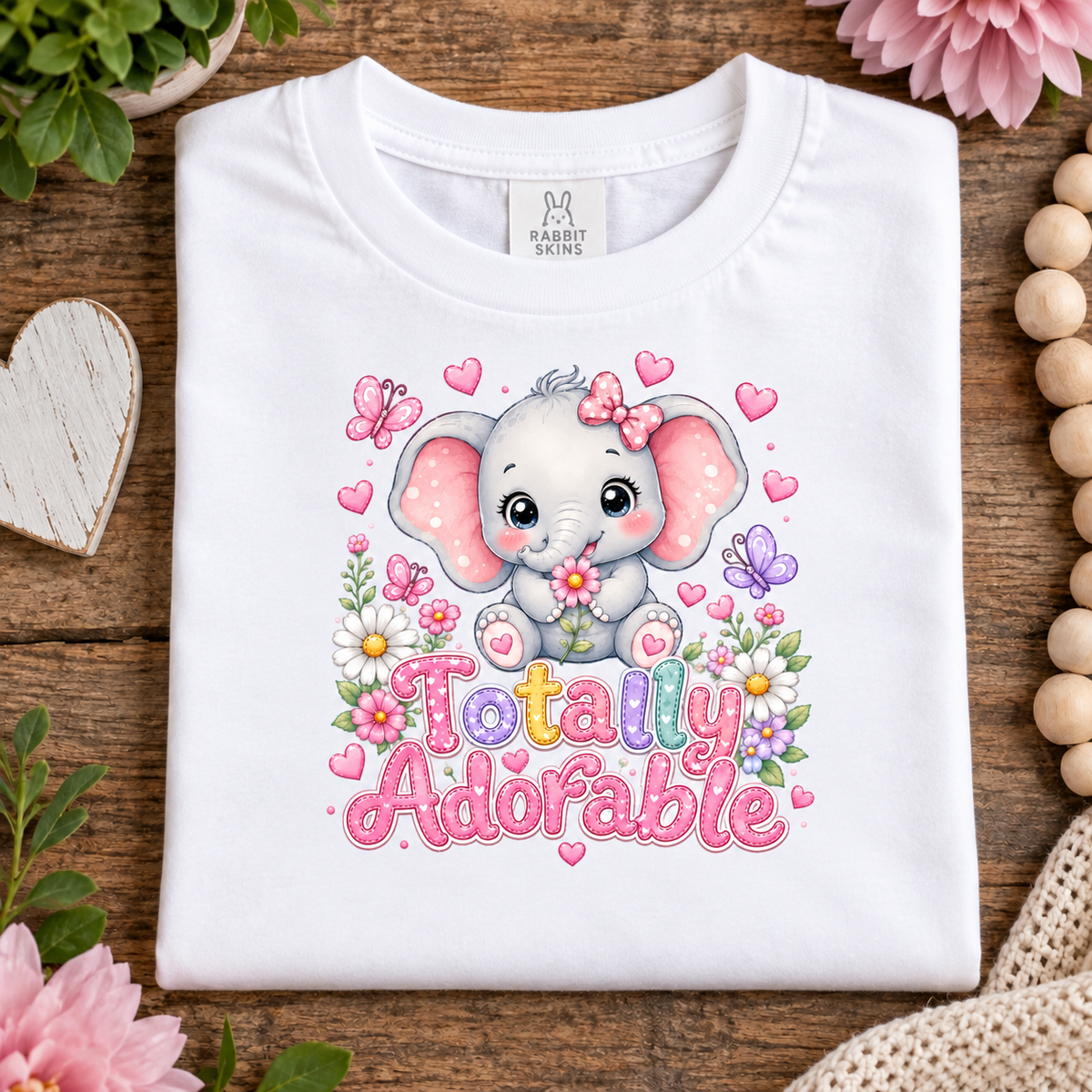A folded white t-shirt with a cute cartoon elephant graphic and the text 'Totally Adorable' in pink letters, styled on a rustic wooden background with decorative flowers and beads.