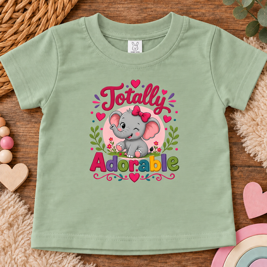 A sage green short-sleeve baby t-shirt with a colorful graphic of a winking cartoon elephant and the text 'Totally Adorable' in pink and multicolor lettering. The t-shirt is displayed flat on a rustic wooden surface surrounded by baby-themed decorative props like a wicker basket, wooden hearts, and a plush rug.
