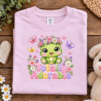 A folded ballerina pink t-shirt with a colorful cartoon graphic of a green character surrounded by flowers, butterflies, and the text 'Totally Adorable', displayed on a rustic wooden background with decorative props.