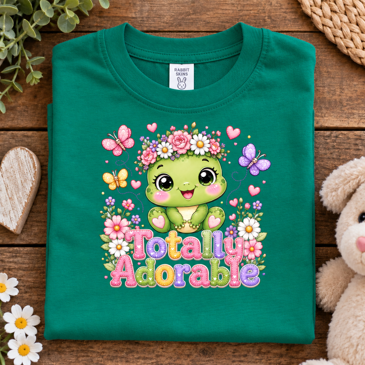 A folded kelly green t-shirt with a colorful cartoon turtle graphic and the text 'Totally Adorable' on a rustic wooden background, styled with a stuffed bunny, daisies, and a heart-shaped wooden block.