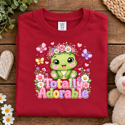 A folded vintage red crewneck t-shirt with a colorful cartoon graphic of a green turtle wearing a flower crown and the text 'Totally Adorable' on a rustic wooden background with a plush toy and flowers.