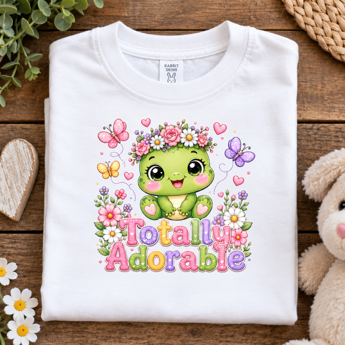 A folded white short-sleeve T-shirt with a colorful cartoon graphic of a green smiling character surrounded by flowers, butterflies, hearts, and the words 'Totally Adorable,' styled on a wooden surface with decorative props.