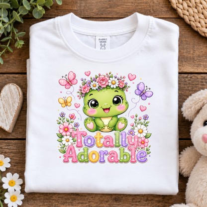 A folded white short-sleeve T-shirt with a colorful cartoon graphic of a green smiling character surrounded by flowers, butterflies, hearts, and the words 'Totally Adorable,' styled on a wooden surface with decorative props.
