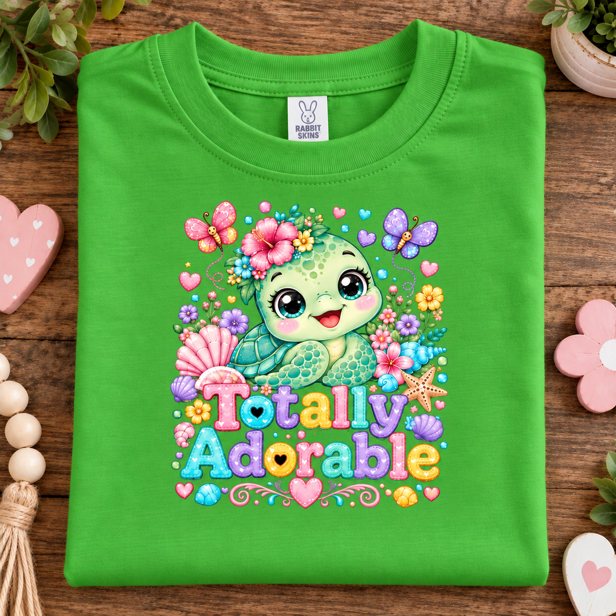 A apple green folded t-shirt with a colorful cartoon turtle graphic and the text 'Totally Adorable' on the front, displayed on a wooden surface with decorative props.