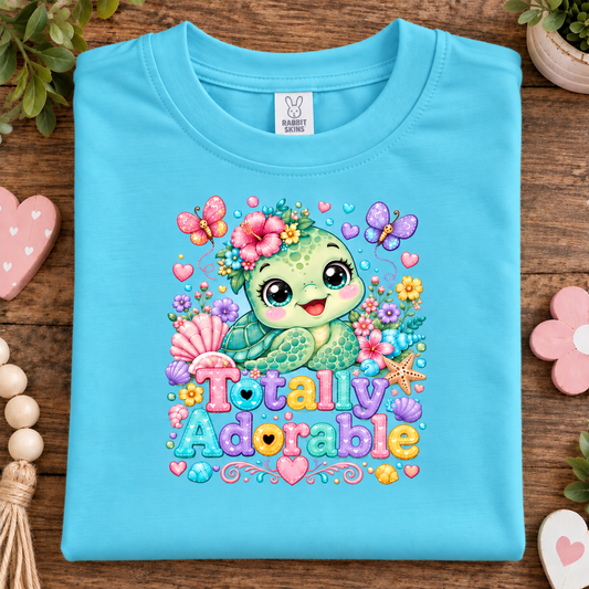 A folded aqua blue t-shirt with a colorful cartoon sea turtle graphic and the text 'Totally Adorable' in pastel letters, displayed on a wooden surface with decorative props.