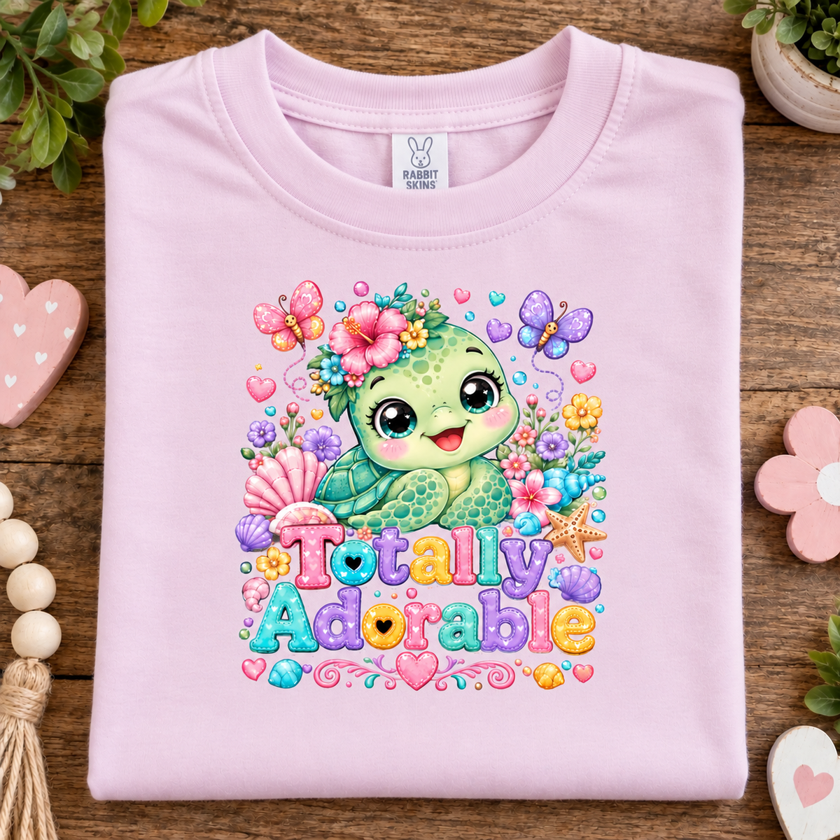 A folded ballerina pink t-shirt with a colorful cartoon turtle graphic and the text 'Totally Adorable' on the front, displayed on a wooden surface with decorative props.