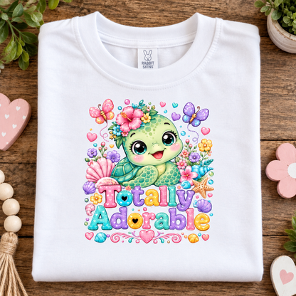 A folded white t-shirt with a colorful cartoon sea turtle graphic and the text 'Totally Adorable' on a wooden surface with decorative props.
