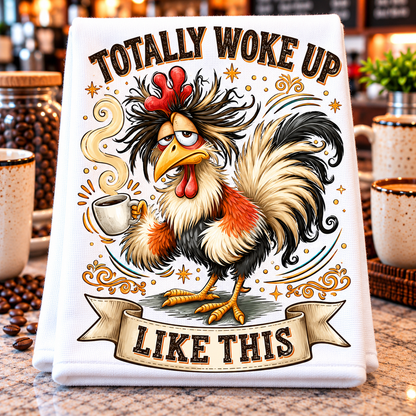 A white kitchen towel or tea towel displayed on a countertop, featuring a colorful graphic of a rooster holding a mug with the text 'TOTALLY WOKE UP LIKE THIS'. The background shows a blurred kitchen setting with coffee beans and mugs.