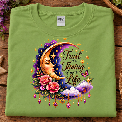 A folded light green (kiwi) Gildan Softstyle T-shirt is displayed on a rustic wooden surface. The front of the shirt features a colorful graphic of a smiling crescent moon face, pink roses, purple clouds, and a butterfly, with the text 'Trust the Timing of your Life' written in a stylized font.