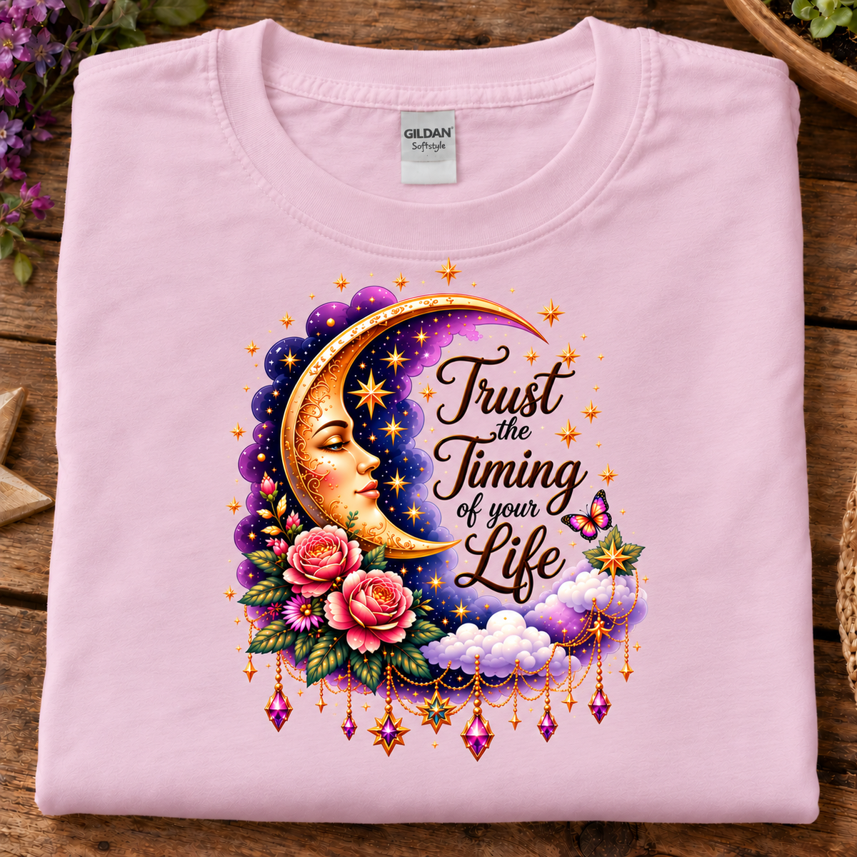 A folded light pink Gildan Softstyle t-shirt is displayed on a rustic wooden surface. The front of the shirt features a detailed graphic print of a crescent moon with a face, pink roses, clouds, and the text 'Trust the Timing of your Life' in a stylized font.