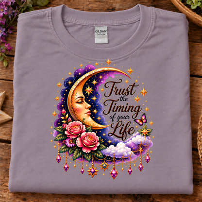 A folded light purple (paragon) Gildan t-shirt displayed flat on a rustic wooden surface, featuring a colorful graphic of a crescent moon, pink roses, clouds, and the text 'Trust the Timing of your Life'.