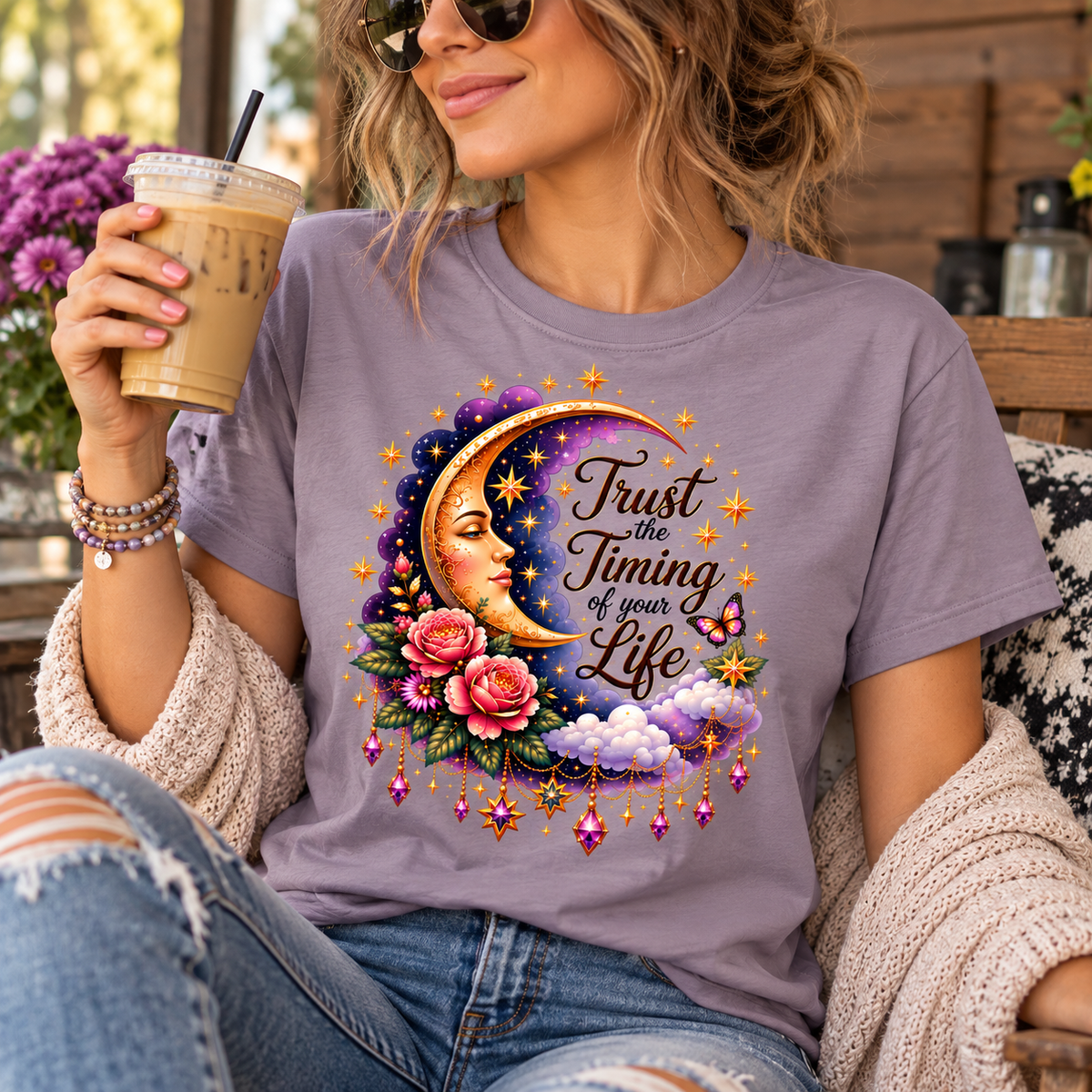 A woman wearing a lavender graphic t-shirt with a colorful moon, flowers, and the text 'Trust the Timing of your Life' printed on the front. She is holding an iced coffee in a cozy outdoor patio setting.