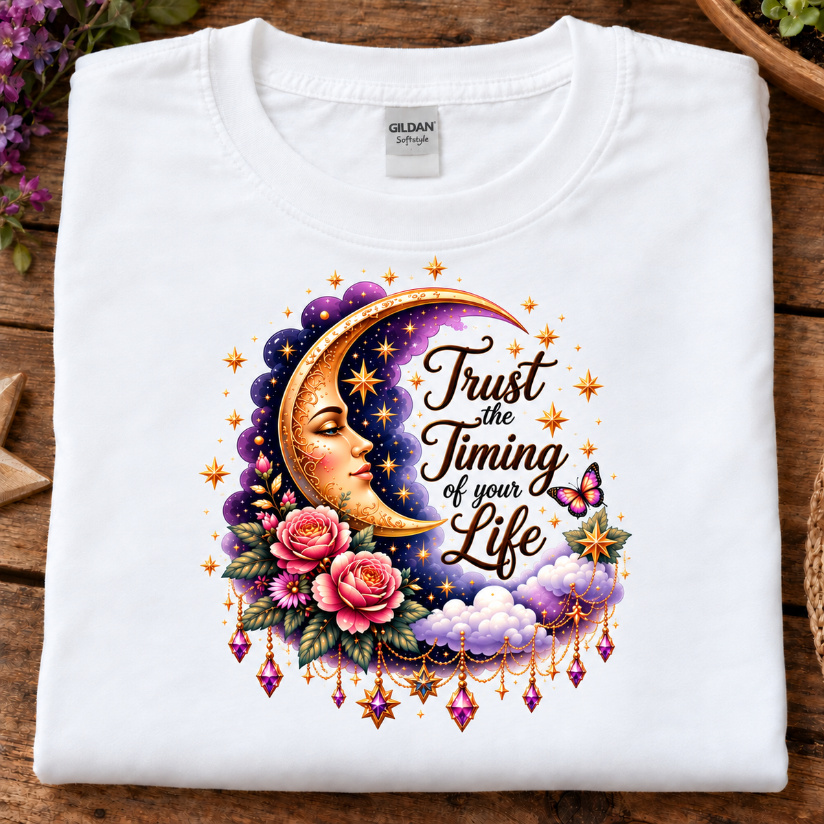 A folded white Gildan t-shirt displayed flat on a rustic wooden surface, featuring a colorful graphic print of a crescent moon face, pink roses, clouds, and the text 'Trust the Timing of your Life'.