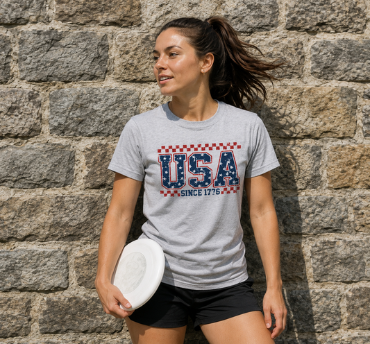 Woman wearing a Sport Grey Gildan SoftStyle patriotic USA Since 1776 graphic t-shirt standing in front of a stone wall holding a frisbee.