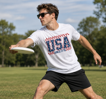 Man wearing a White Gildan SoftStyle patriotic USA Since 1776 graphic t-shirt standing in front of a stone wall holding a frisbee.