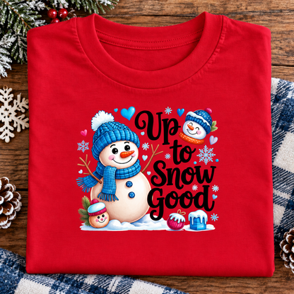 A folded red crewneck t-shirt is displayed on a rustic wooden surface with holiday decorations. The front of the shirt features a colorful graphic of snowmen wearing blue and orange winter hats and scarves, surrounded by snowflakes, hearts, and the text 'Up to Snow Good'.