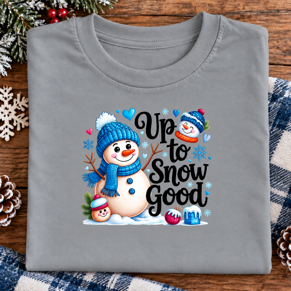 A folded gray t-shirt with a colorful 'Up to Snow Good' winter graphic, displayed on a wooden surface with holiday decorations like pinecones, a snowflake, and a plaid blanket.