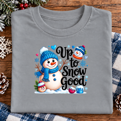 A folded gray t-shirt with a colorful 'Up to Snow Good' winter graphic, displayed on a wooden surface with holiday decorations like pinecones, a snowflake, and a plaid blanket.