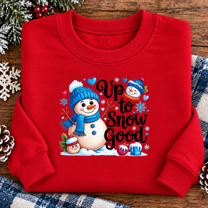 A red crewneck sweatshirt with a festive 'Up to Snow Good' graphic featuring cartoon snowmen, blue hats, and snowflakes, laid flat on a rustic wooden background with holiday decorations.