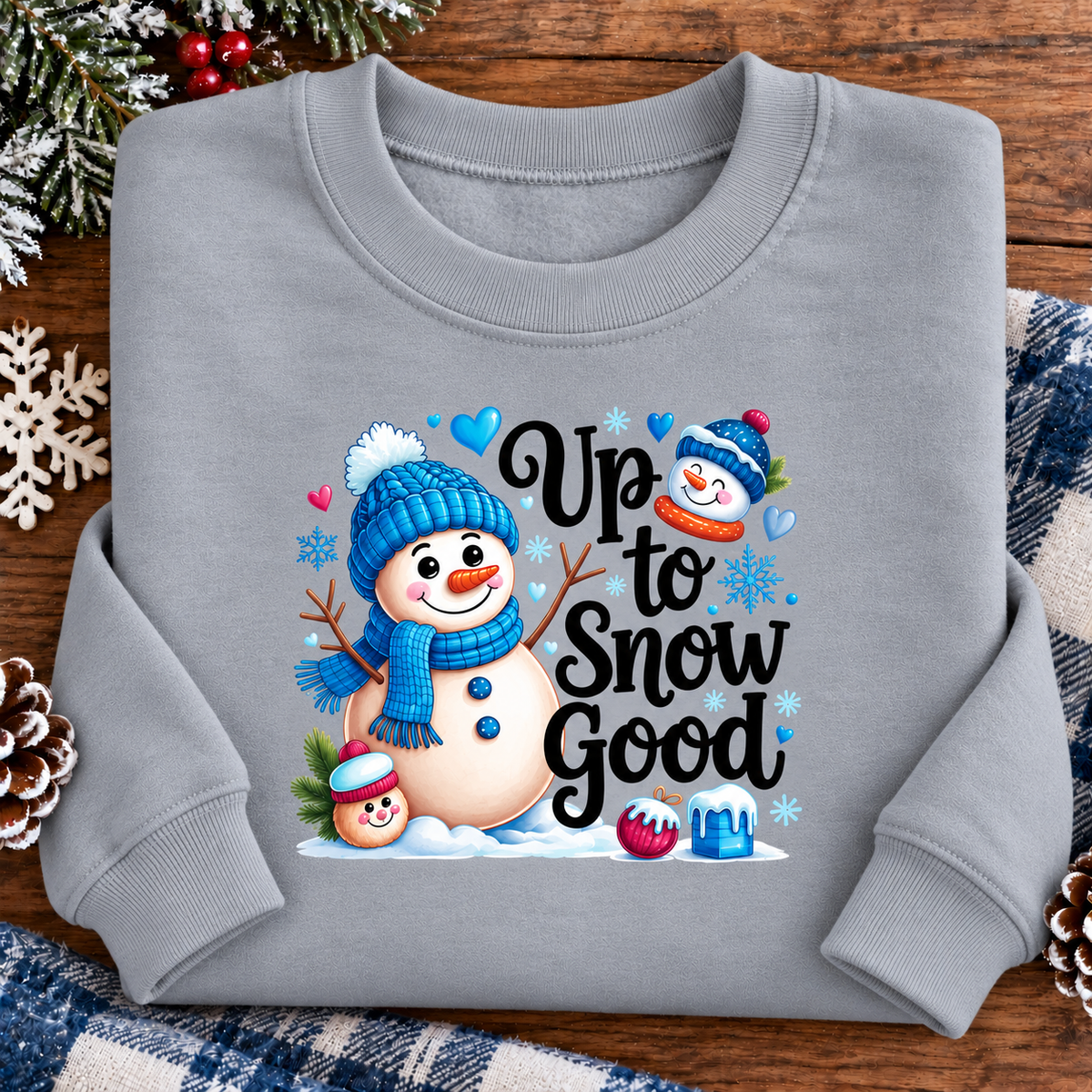 A light gray crewneck sweatshirt featuring a festive Christmas graphic with snowmen, hearts, and the text 'Up to Snow Good'. The sweatshirt is displayed flat on a rustic wooden surface surrounded by holiday decorations like pinecones, a snowflake, and a plaid blanket.