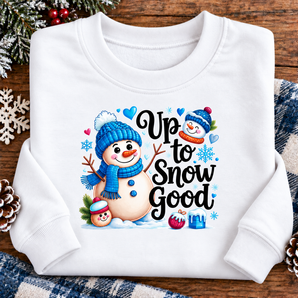 A white long-sleeve crewneck sweatshirt with a colorful winter graphic that reads 'Up to Snow Good' alongside cartoon snowmen, hearts, and snowflakes, displayed on a rustic wooden background with holiday decorations.
