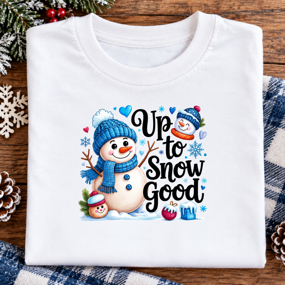A folded white crewneck t-shirt with a colorful winter graphic that reads 'Up to Snow Good' featuring cartoon snowmen, hearts, and snowflakes, displayed on a rustic wooden background with holiday decorations.