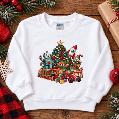 A white Gildan sweatshirt laid flat on a rustic wooden surface, decorated with Christmas-themed props. The sweatshirt features a colorful graphic of toys and a Christmas tree on the front.